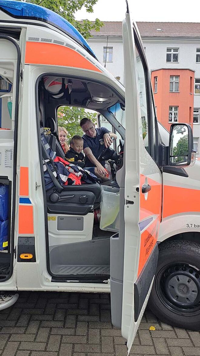 Ein Kind sitzt hinter dem Lenkrad eines Rettungswagens. Zwei junge Menschen in Rettungsdienstkleidung erklären dem Kind, was es dort sieht. Das ist Teil eines Ausbildungsprojektes für angehende Notfallsanitäter.